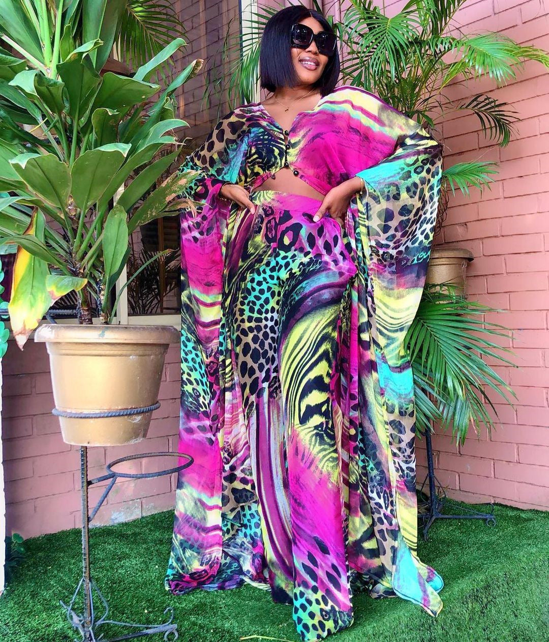 Woman in a colorful outfit standing in front of a pink wall with plants.