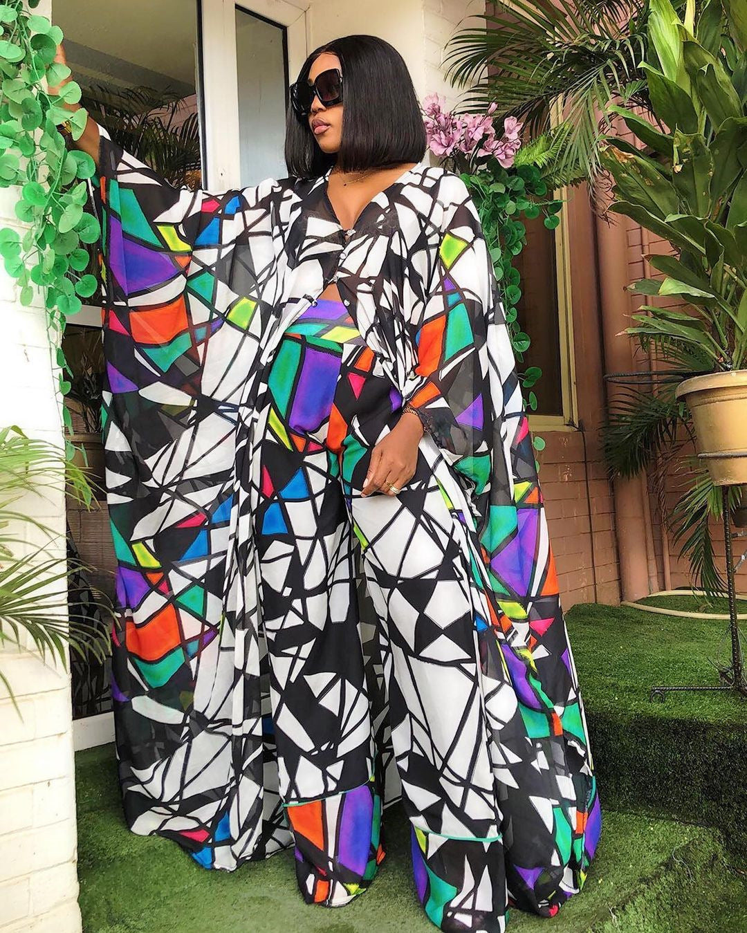 Woman wearing a colorful geometric patterned outfit standing outdoors with plants around.