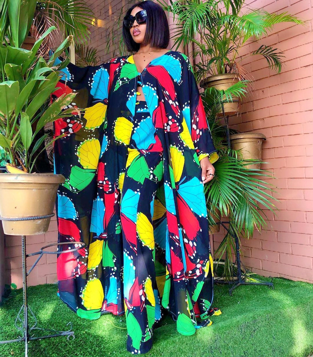 Woman wearing a colorful patterned outfit standing in front of potted plants.