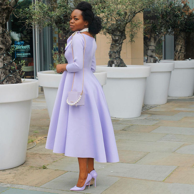 Woman in a lavender dress standing outdoors with large white planters and trees in the background.