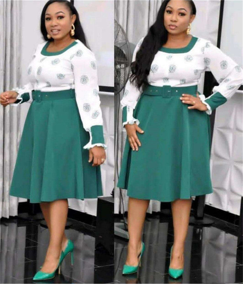 Woman wearing a white blouse with green patterns and a green skirt, standing against a white curtain background.