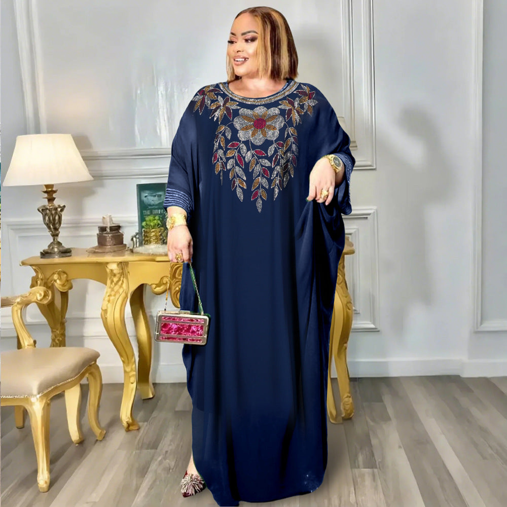 Woman in a long dark blue dress with floral embroidery standing in a room with a gold table and chair.