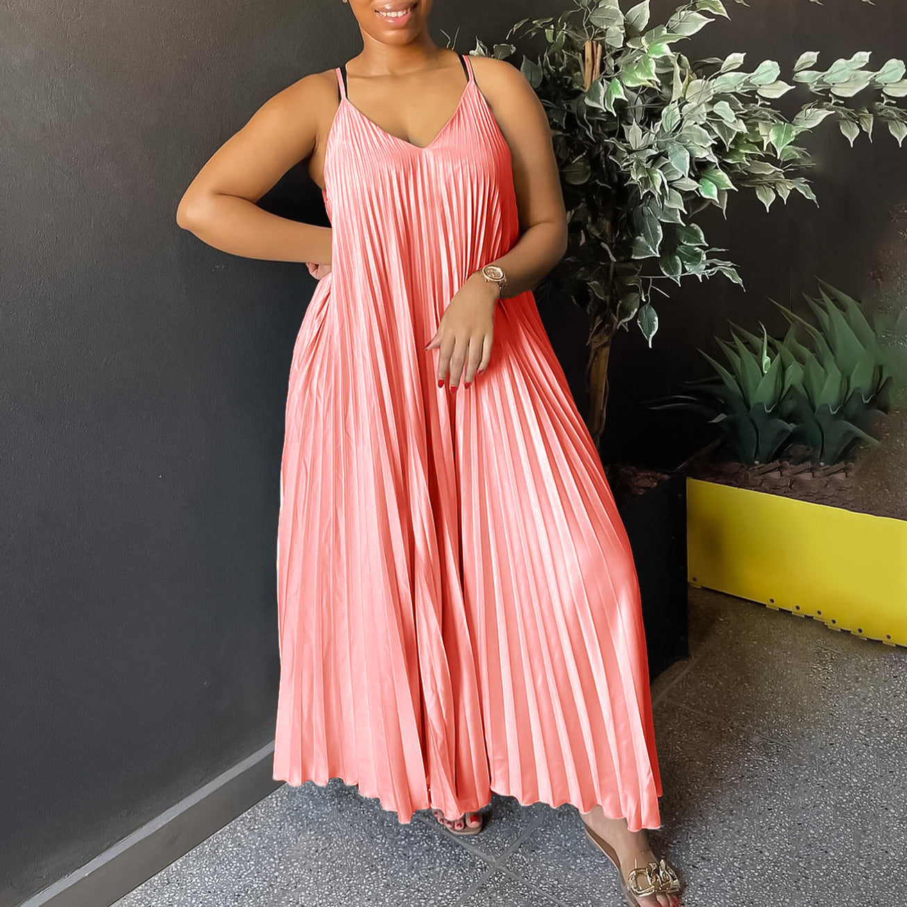 Woman wearing a pink pleated dress standing against a dark wall with plants.