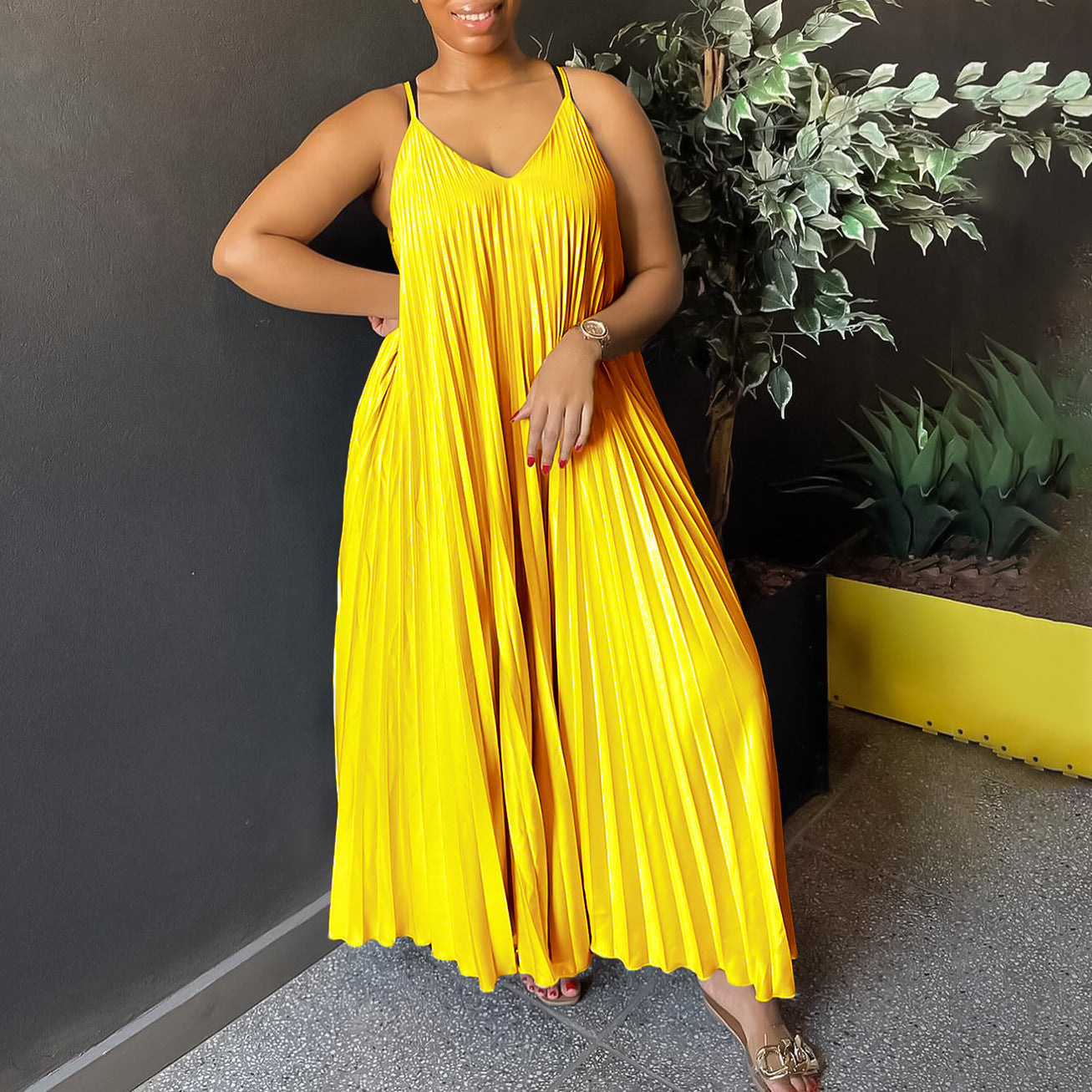 Woman wearing a yellow dress standing against a dark wall with plants.
