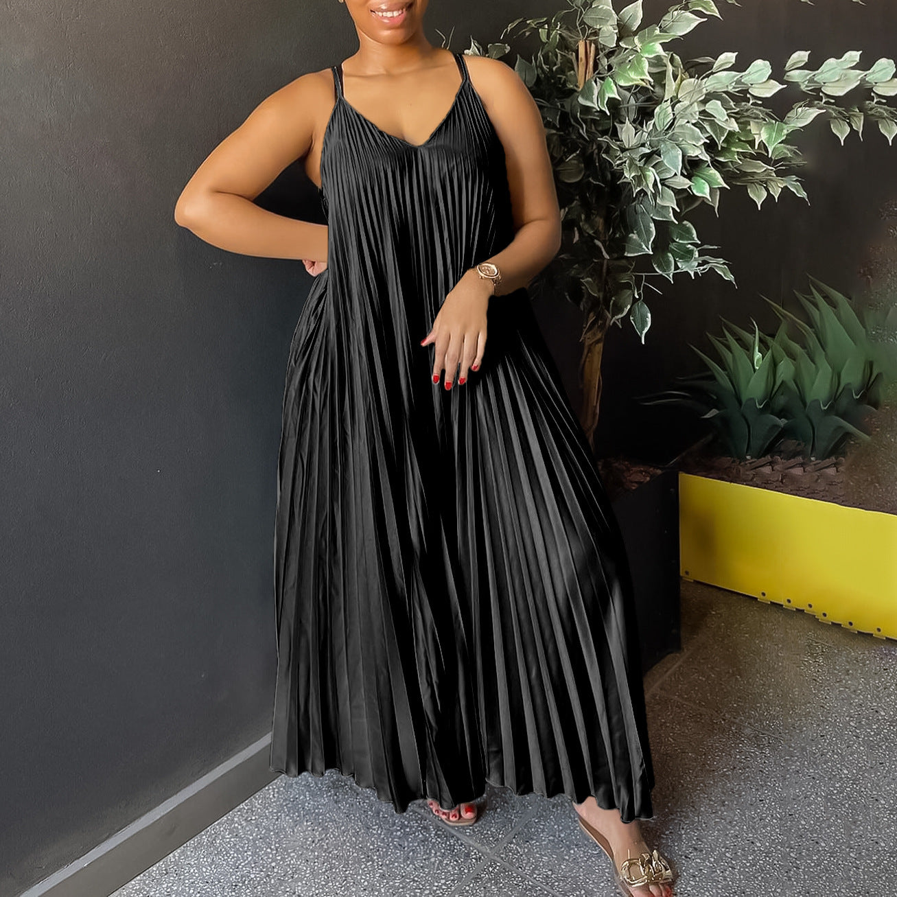 Woman wearing a black pleated dress standing against a dark wall with plants.