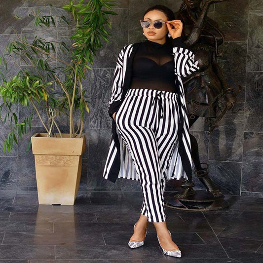 Woman in black and white striped outfit standing against a dark stone wall with a plant.