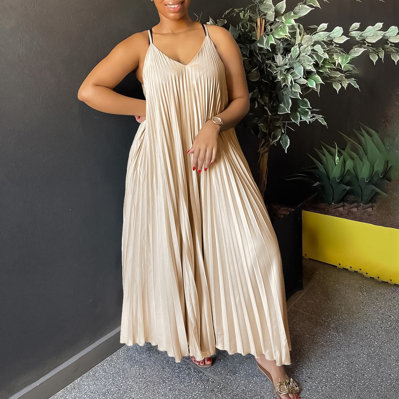 Woman wearing a beige pleated dress standing against a dark wall with plants.