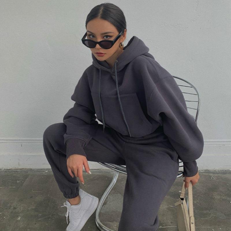 Person wearing a dark gray hoodie and pants sitting on a chair against a plain wall.