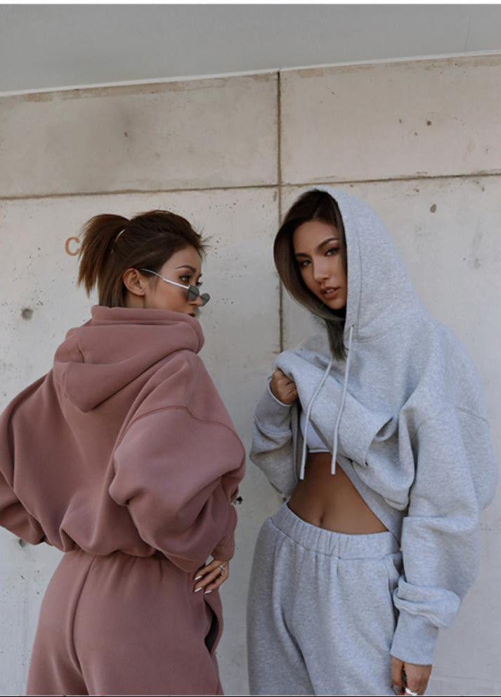 Two women wearing matching hoodie and pants outfits against a concrete wall.
